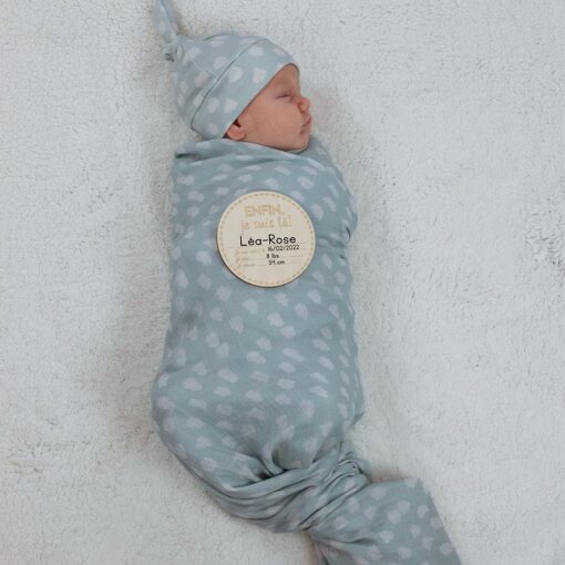 bébé emmailloté dans une mousseline et bonnet bleu avec pastille de bois montrant ses informations de naissance