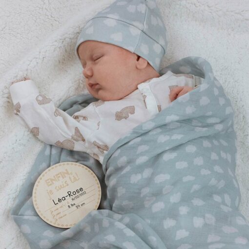 bébé dort dans une mousseline et bonnet bleu avec pastille de bois montrant ses informations de naissance