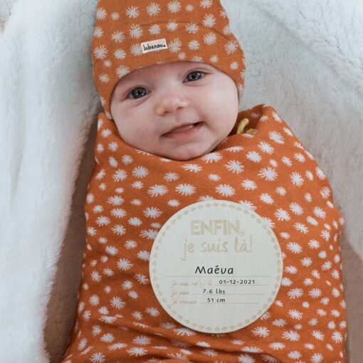 Petite fille emmaillotée dans un ensemble orange avec pastille de bois qui annonce détails de naissance de bébé