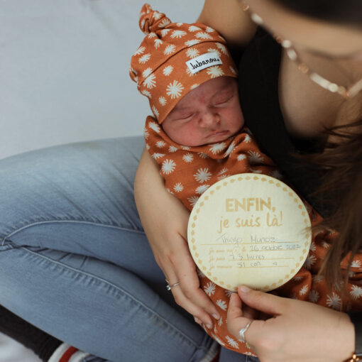 Nouveau-né emmailloté dans un ensemble orange dans les bras de maman qui tient pastille de bois montrant les informations de naissance de son bébé vue de haut
